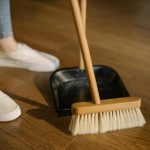 Close-up of cleaning process with broom and dustpan beside sneakers on a wooden indoor floor.